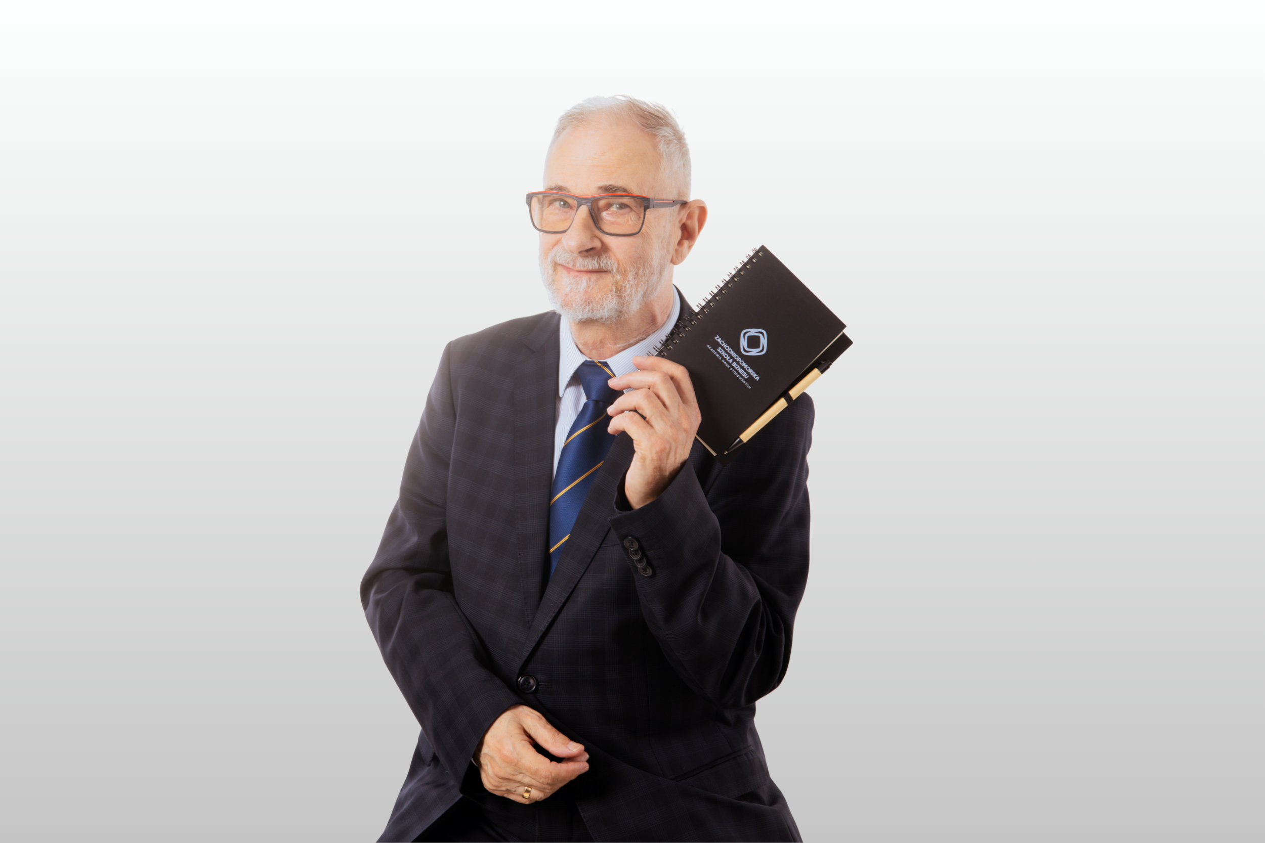 Starszy mężczyzna w garniturze i okularach uśmiecha się, trzymając czarny spiralny notatnik z dołączonym długopisem, na jasnym tle.