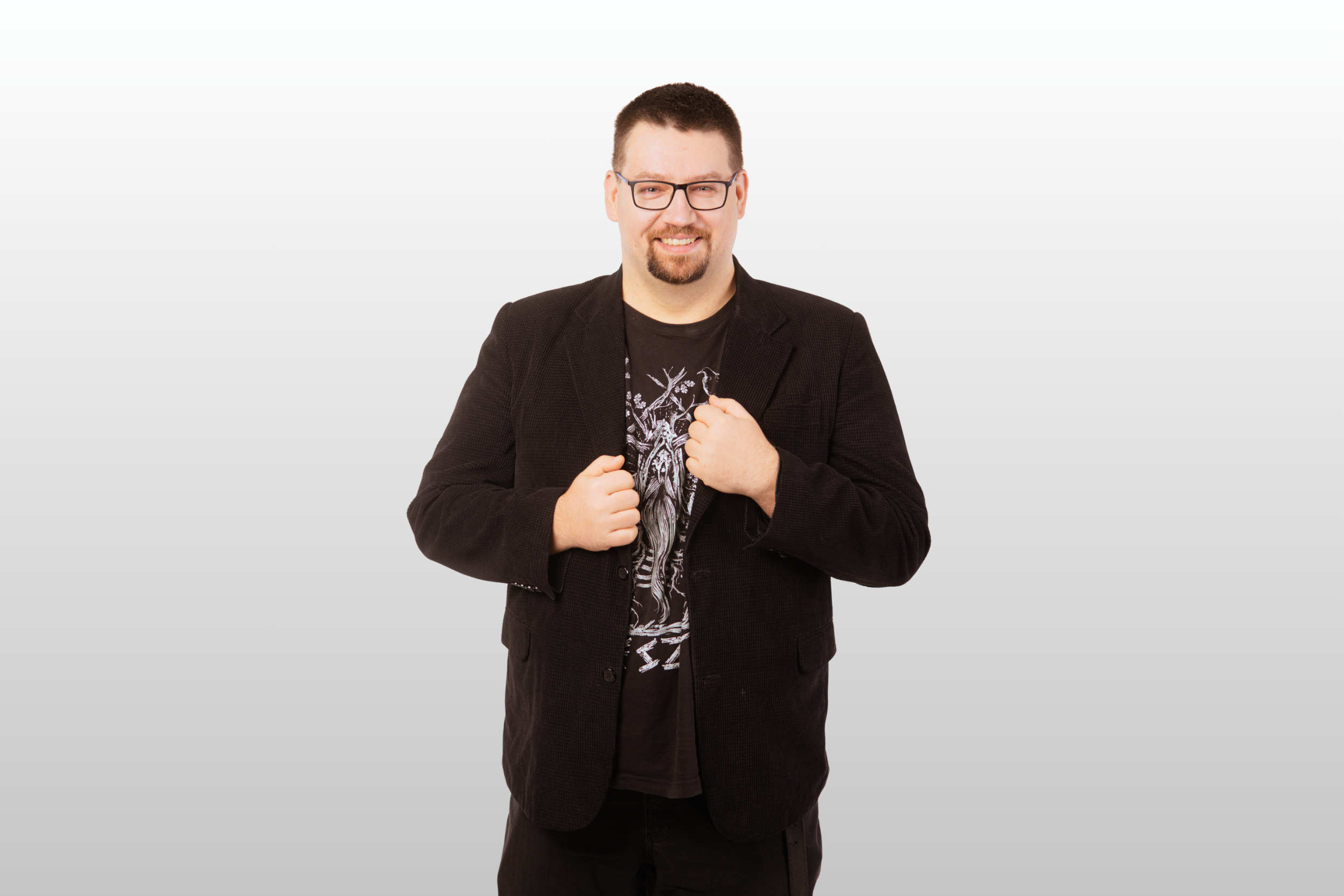 Mężczyzna z krótkimi ciemnymi włosami, okularami i brodą uśmiecha się, ubrany w czarną marynarkę na koszulce z grafiką, stojąc na prostym białym tle.