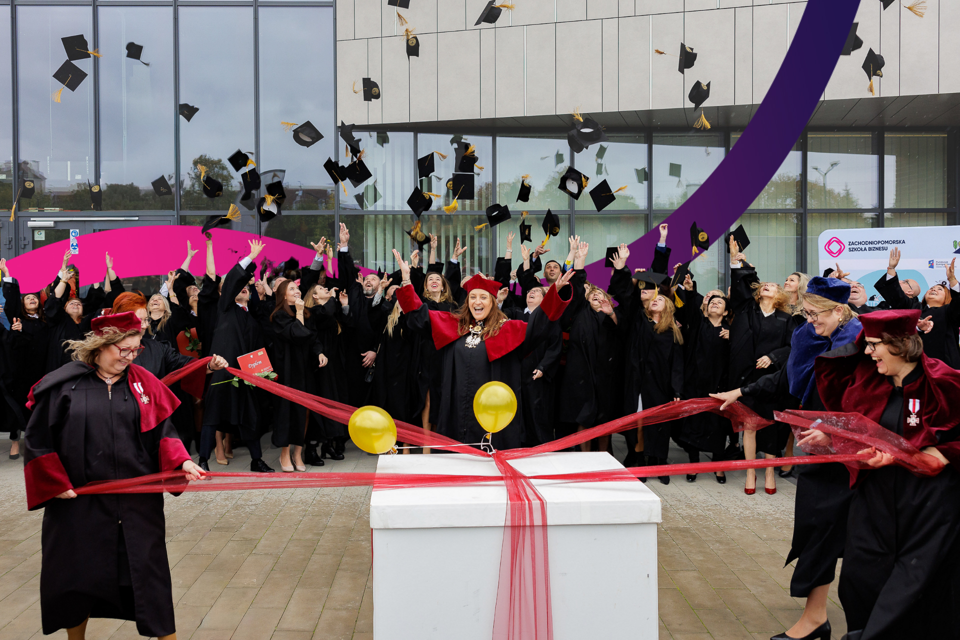Duża grupa absolwentów w czarnych togach i czapkach świętuje na świeżym powietrzu, wyrzucając czapki w powietrze. Rektorzy i dziekani w czerwono-czarnych szatach stoją w pobliżu wstążki i balonów, rozpakowują wielki prezent, a w tle widać nowoczesny budynek.