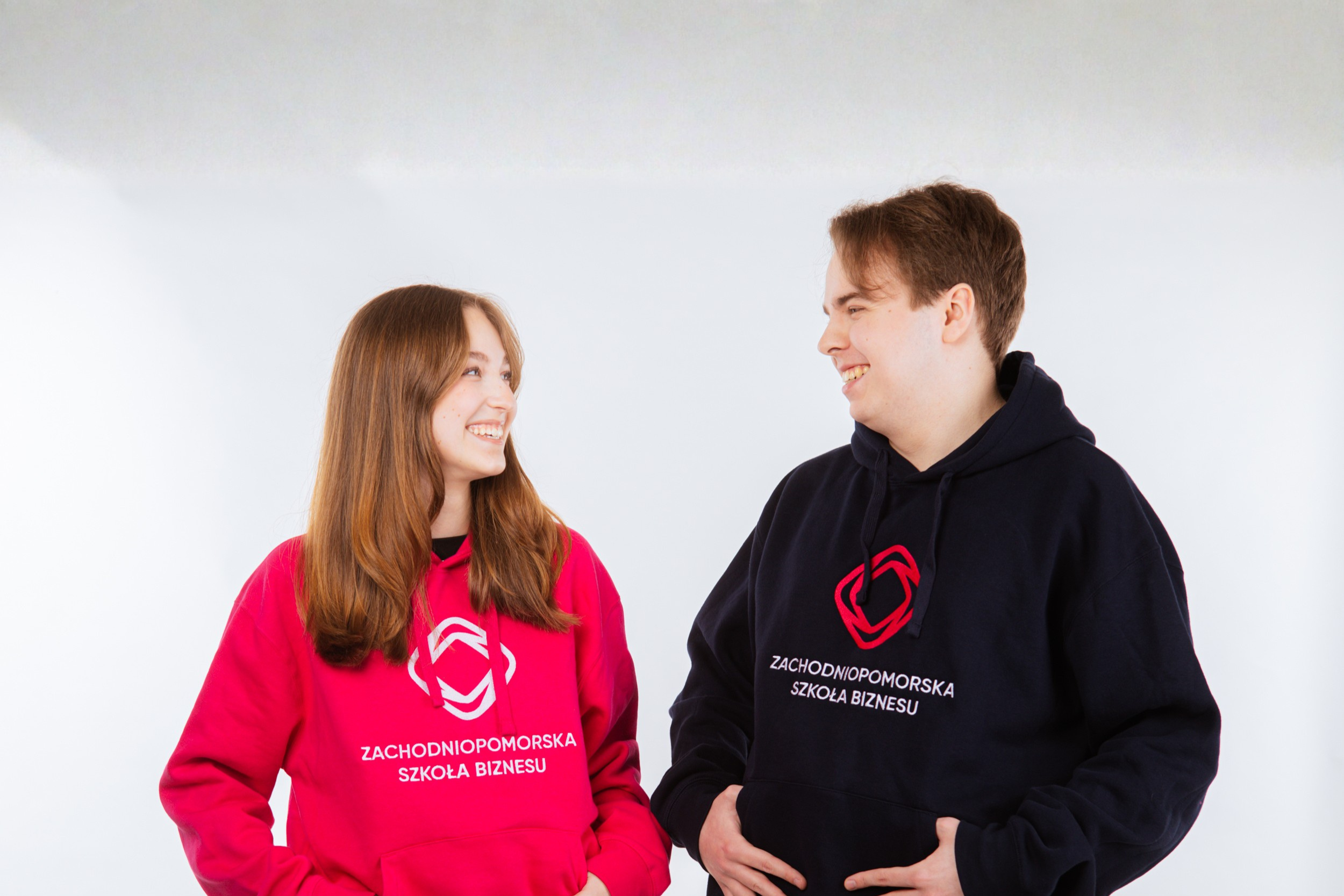 Dwoje młodych ludzi, dziewczyna i chłopak, stoi naprzeciwko siebie, uśmiechnięci, ubrani w bluzy z logo i napisem Zachodniopomorska Szkoła Biznesu. Dziewczyna ma na sobie jasnoróżową bluzę z kapturem, chłopak granatową, obaj pozują z rękami na biodrach