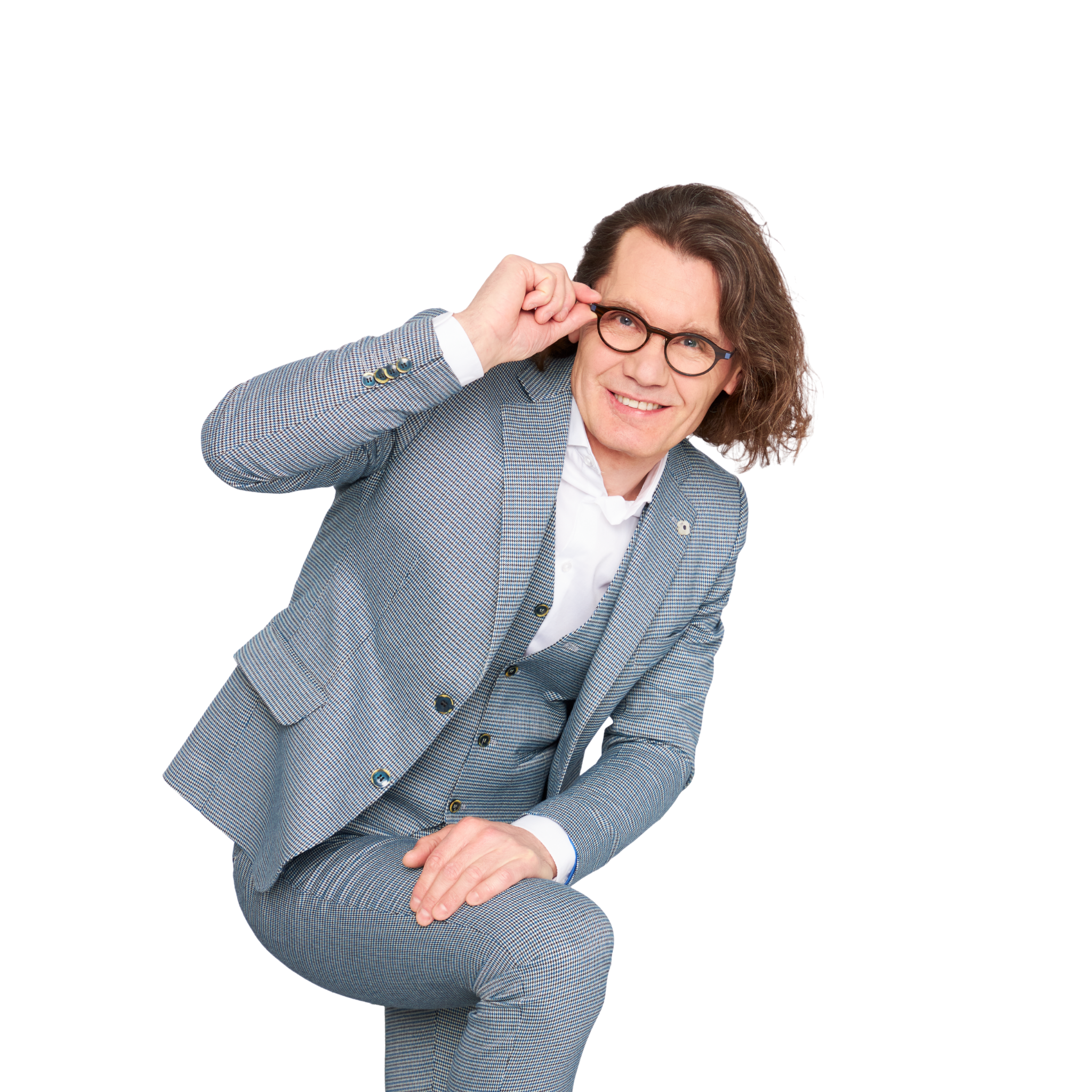 Uśmiechnięty mężczyzna w średnim wieku, w szarej marynarce, opiera się o zgięte kolano i prawą ręką sięga do okularów.