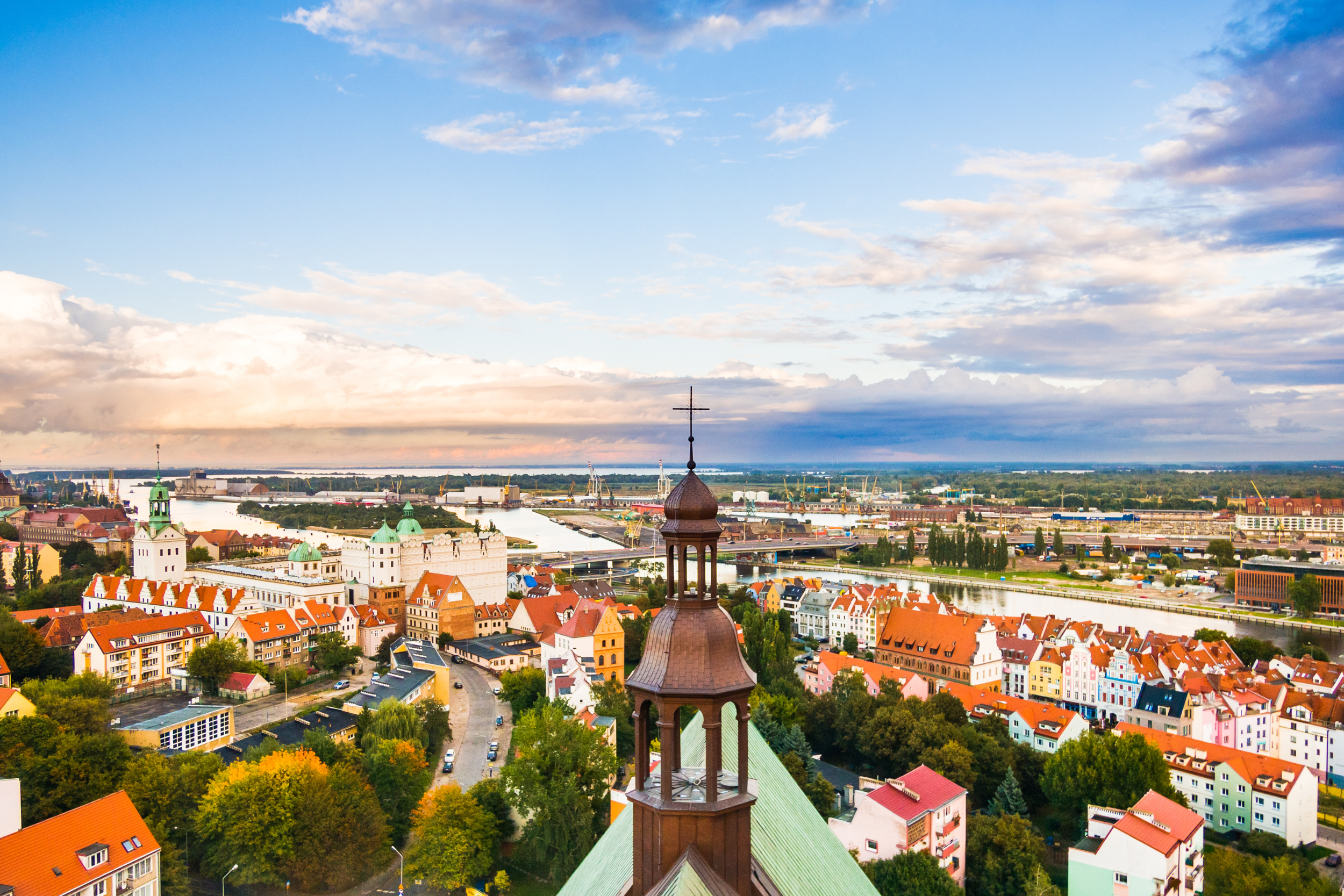 Malowniczy pejzaż miejski z centralną wieżą kościoła, kolorowymi budynkami z dachami pokrytymi czerwoną dachówką, krętymi drogami, zielonymi drzewami i rzeką w tle pod częściowo zachmurzonym niebem.