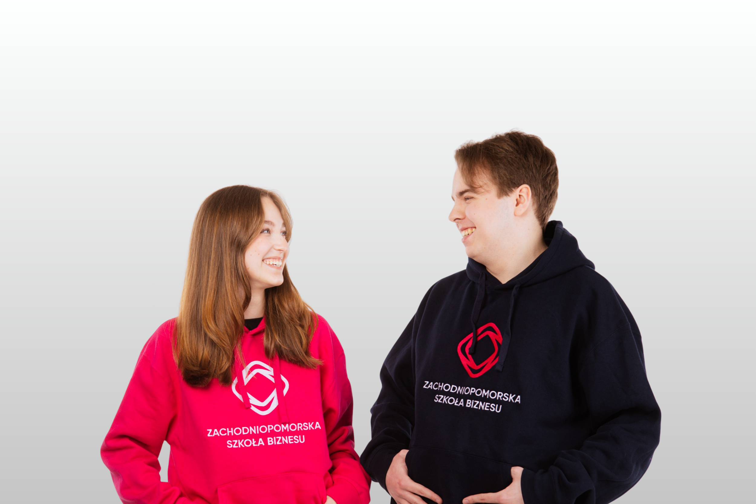 Dwoje młodych ludzi, dziewczyna i chłopak, stoi naprzeciwko siebie, uśmiechnięci, ubrani w bluzy z logo i napisem Zachodniopomorska Szkoła Biznesu. Dziewczyna ma na sobie jasnoróżową bluzę z kapturem, chłopak granatową, obaj pozują z rękami na biodrach
