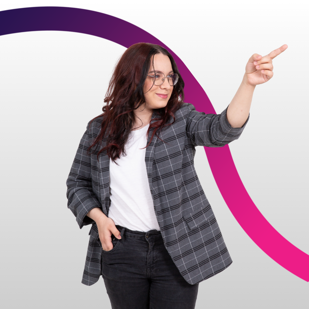 Młoda dziewczyna, ubrana w marynarkę w kratę i czarne dżinsy patrzy się w lewo i pokazuje coś palcem