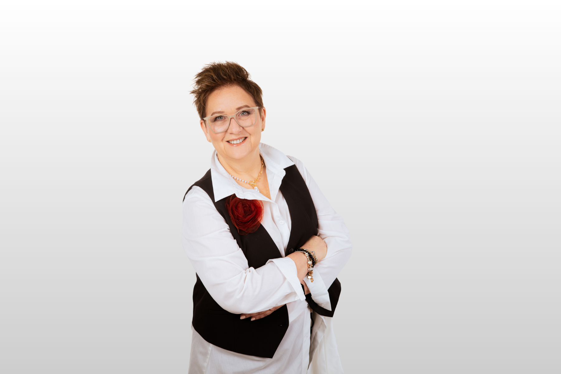 Dojrzała kobieta w krótkich rudych włosach, ubrana elegancko w koszulę i czarną kamizelkę, stoi uśmiechnięta z założonymi rękami