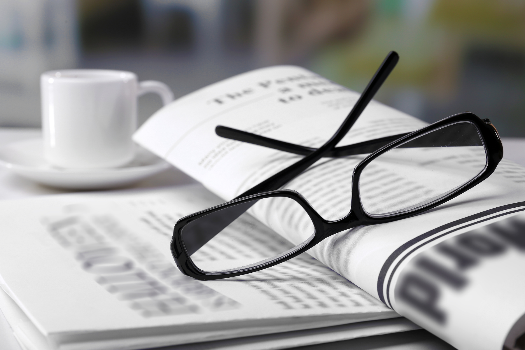 Na gazetach leżą czarne okulary korekcyjne, w tle stoi biała filiżanka