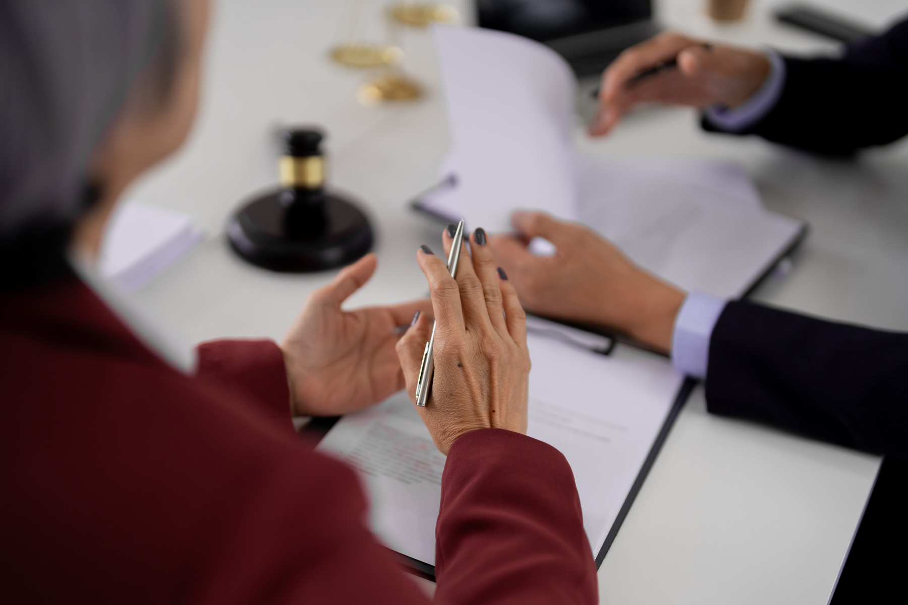 na zdjęciu widać ręce dwóch postaci przy biurku, jedna jest kobietą trzyma srebrny długopis przed soba ma dokumenty, w tle legancką czanrno złotą pieczęć, na dalszym planie widać ręce mężczyzny, widoczne są mankiety od garnituru, trzyma ona dokumenty, 