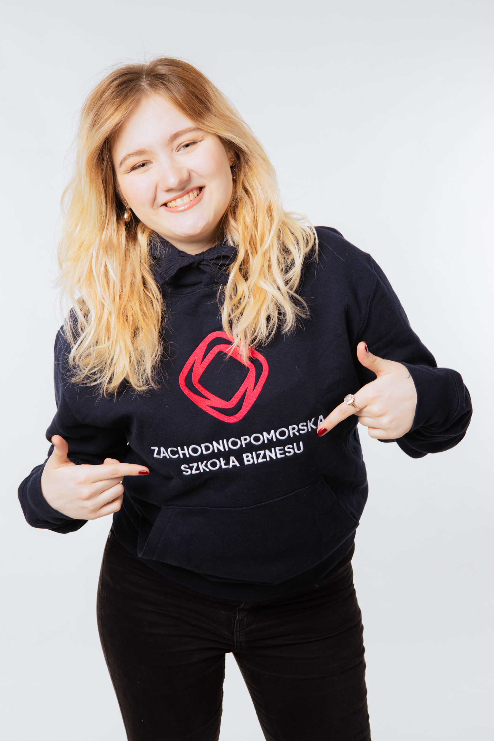 Młoda, uśmiechnięta dziewczyna, blondynka, stoi w granatowej bluzie z logo Zachodniopomorskiej Szkoły Biznesu i wskazuje na logo palcami