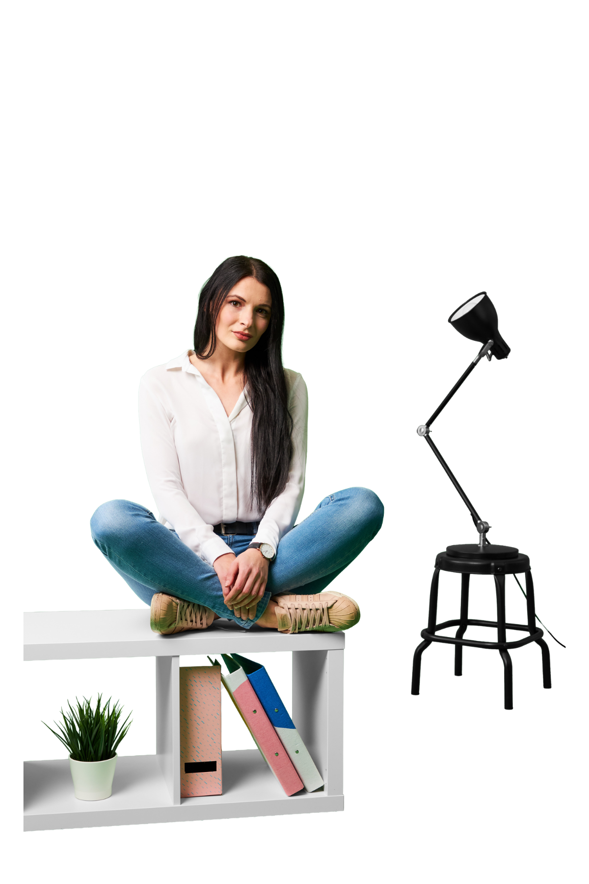 na białej szafce siedzi młoda studentka z długimi czarnymi włosami, ze skrzyżówanymi nogami i lekko się uśmiecha, z tle widać czarną lampkę stojącą na małym, czarnym taborecie. 