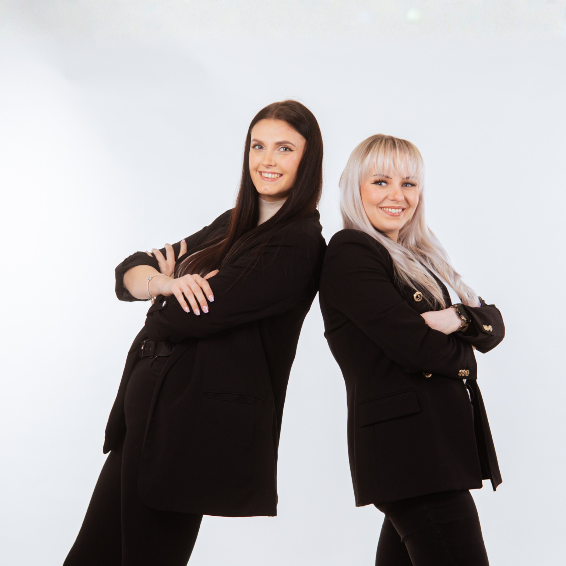 dwie uśmiechnięte dziewczyny, brunetka i blondynka, ubrane w czarne marynarki stoją oparte o siebie plecami z założonymi rękoma