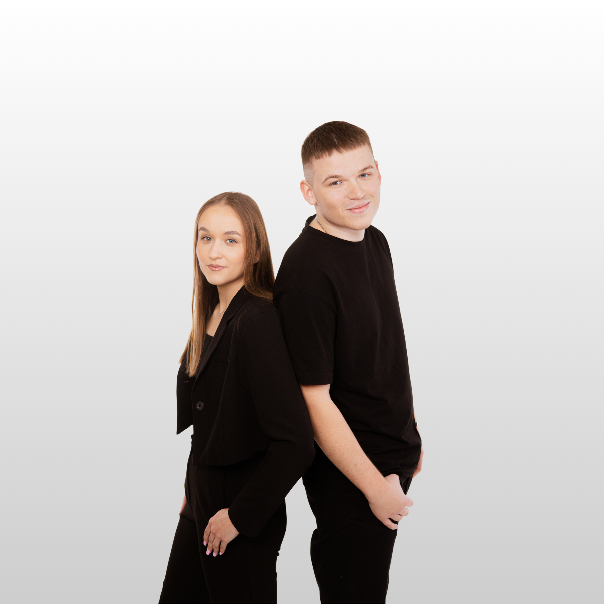 Młoda dziewczyna ubrana na czarnu, blondynka z długimi włosami, stoi tyłem opira się o plecy chłopaka z krótkimi włosami, oboje są ubrani na czarno i patrzą się na wprost lekko uśmiechając się, ręce mają w kieszeni