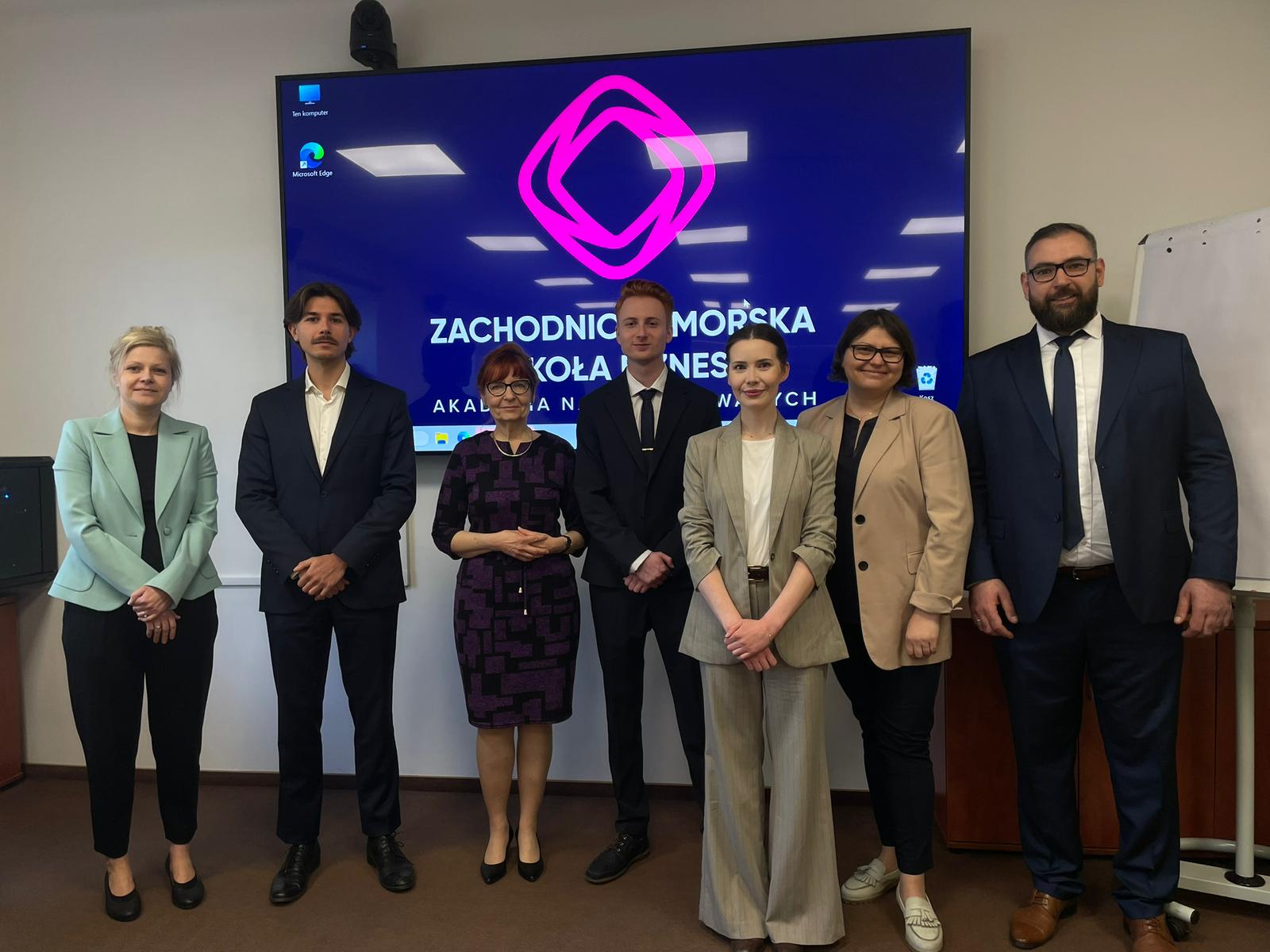 Studenci po obronie pozują do zdjęcia z komisją na tle logo ZPSB