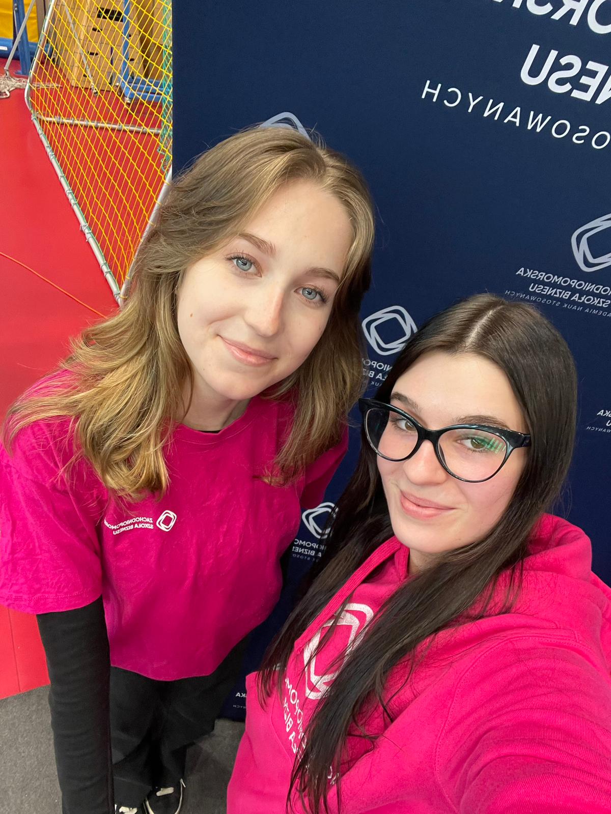 Dwie studentki w różowych ciuchach pozują do zdjęcia na ściance reklamowej