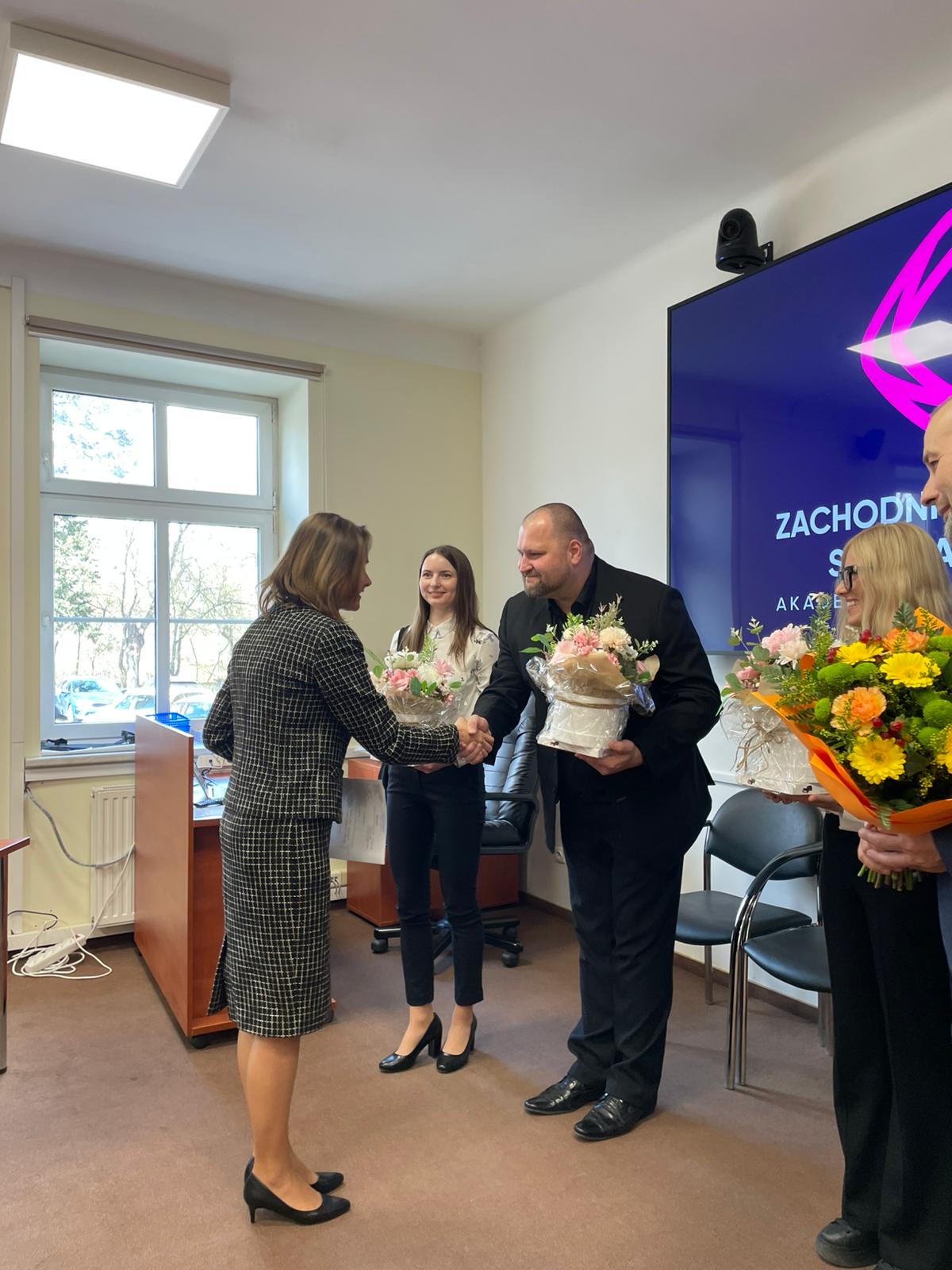 Dziekan Wydziału podaje rękę absolwentowi, który trzyma kwiaty