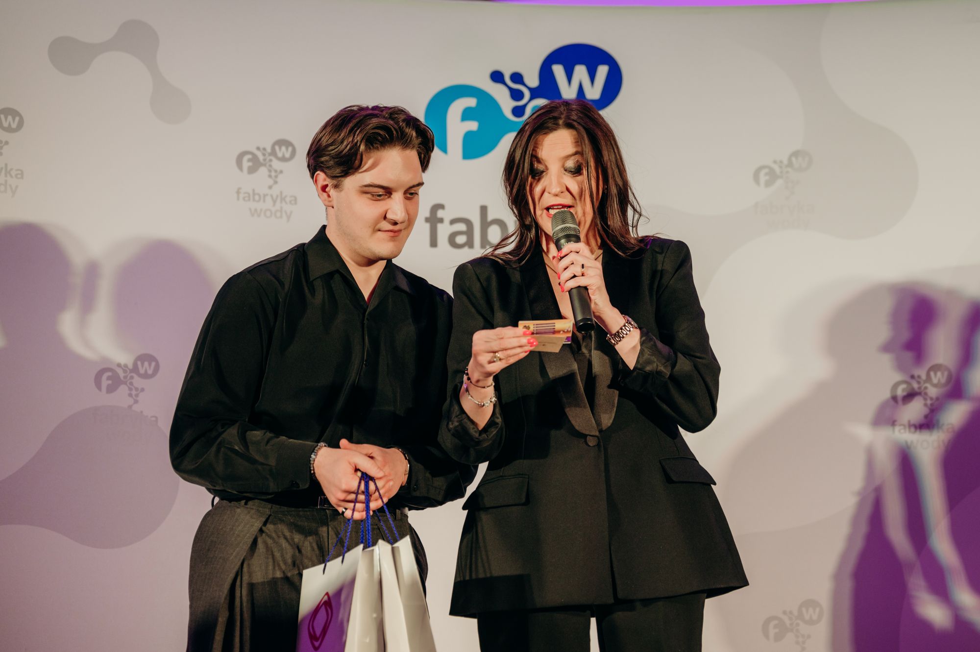 Kobieta w czarnym garniturze przemawia do mikrofonu, trzymając kartkę, obok stoi młody mężczyzna z torbami prezentowymi; za nimi ścianka z logo wydarzenia.