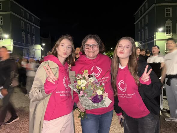 Trzy kobiety pozują do zdjęcia podczas nocnego wydarzenia plenerowego. Dwie pokazują znak pokoju, a środkowa osoba trzyma bukiet kwiatów.