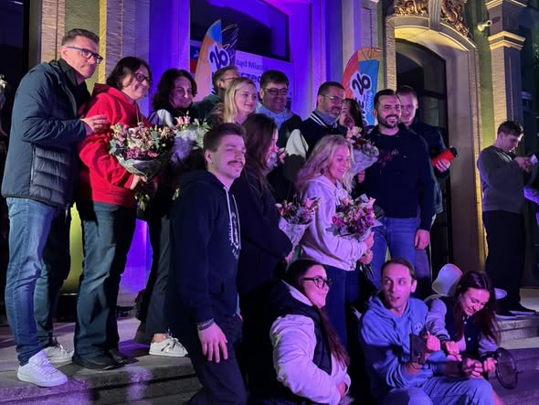 Grupa osób pozuje na schodach przed podświetlonym budynkiem podczas wieczornego wydarzenia.