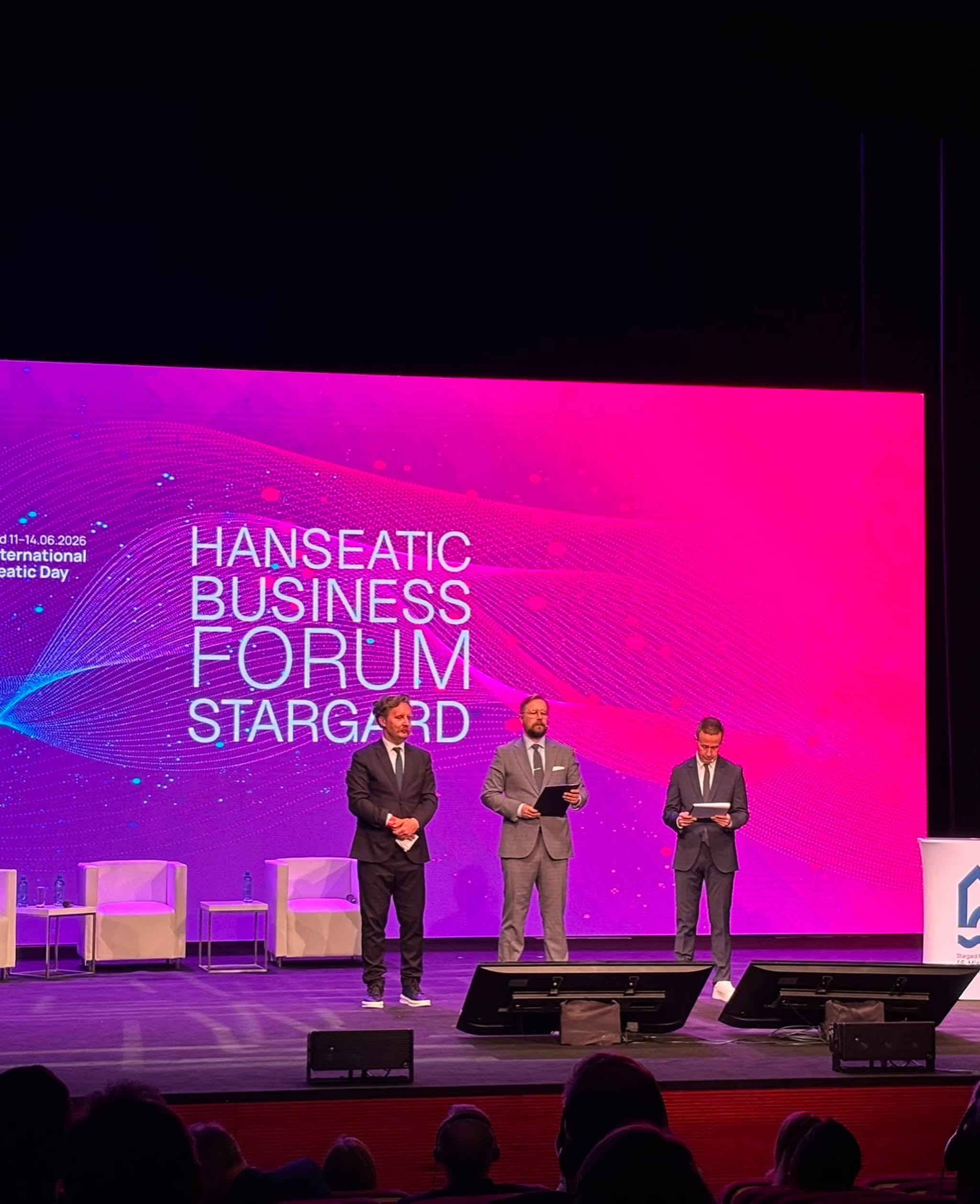 Trzej mężczyźni w garniturach stoją na scenie przed dużym, różowo-fioletowym ekranem z napisem „HANSEATIC BUSINESS FORUM STARGARD”. Mężczyzna w środku trzyma podkładkę z dokumentami i przemawia do mikrofonu. W tle widoczne są białe fotele i stoliki.