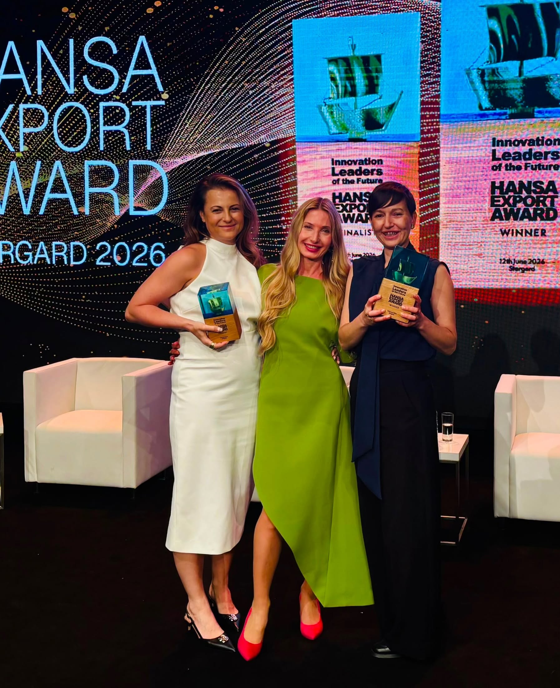 Trzy uśmiechnięte kobiety pozują wspólnie na tle ekranu z napisem „HANSA EXPORT AWARD STARGARD 2026”. Kobiety po bokach trzymają szklano-drewniane statuetki. Kobieta po lewej jest w białej sukience, w środku w limonkowej asymetrycznej kreacji, a po prawej w ciemnym kombinezonie.