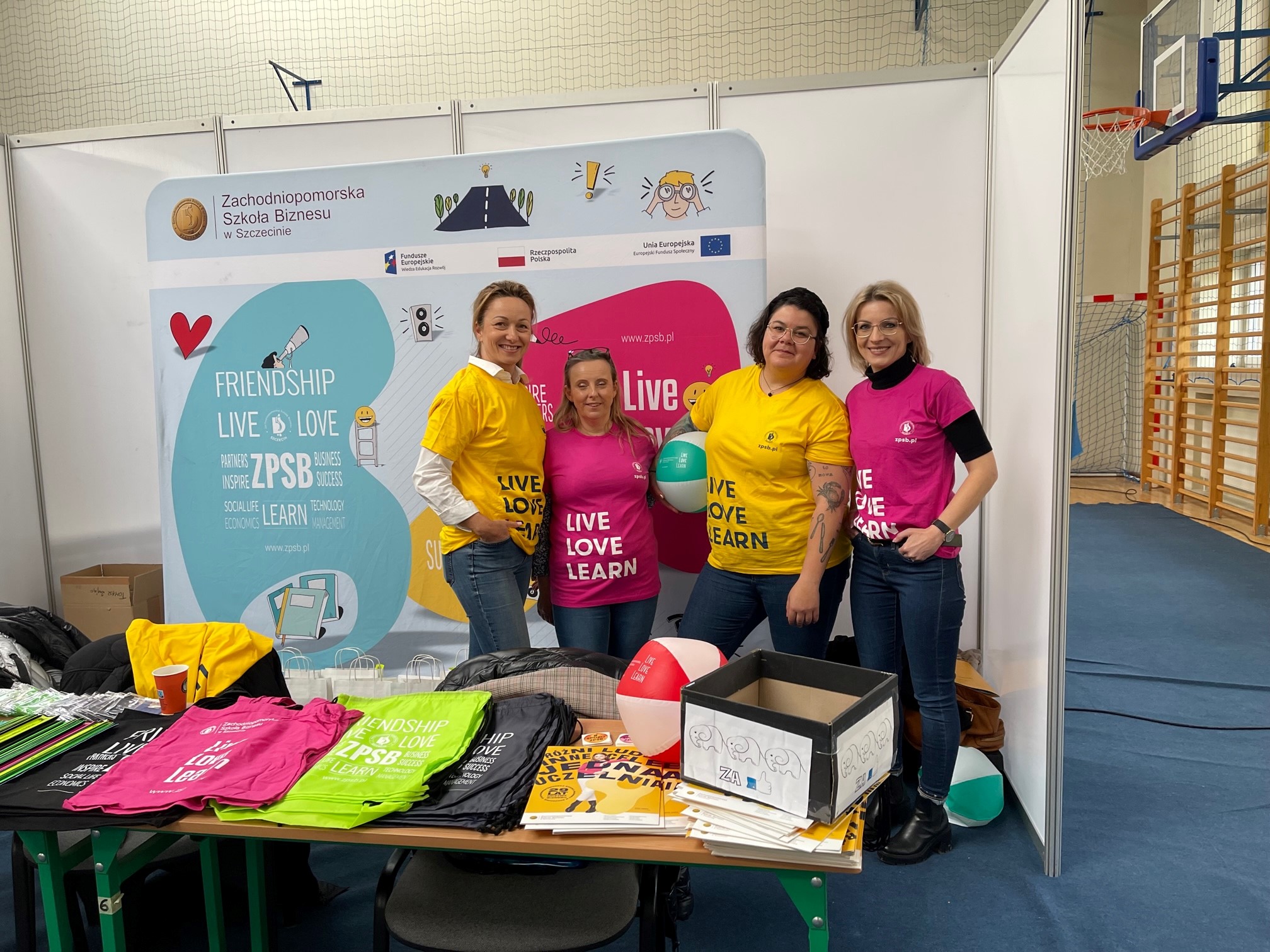 Ilustracja przedstawia cztery kobiety ubrane w żółte i różowe koszulki. Przed nimi znajduje się stolik zastawiony różnymi gadżetami.